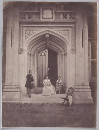 Man standing, hat in hand,  on steps of doorway of Château de Vallombrosa, Cannes with seated woman and girl in front of door. A wooden folding chair stands before the doorway