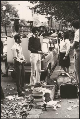 A group of students stand on the curb by piles of books 