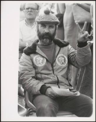 A man in bobbled flat cap holds up two fingers in the peace sign