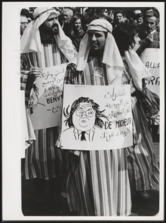 A protest with people dressing up in Arab-style robes, holding placards, some written in Arabic,  the politician De Michelis is illustrated on another