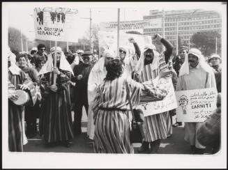 A protest with people dressing up in Arab-style robes, holding placards, some written in Arabic,  the politician Pierre Carniti is illustrated on another