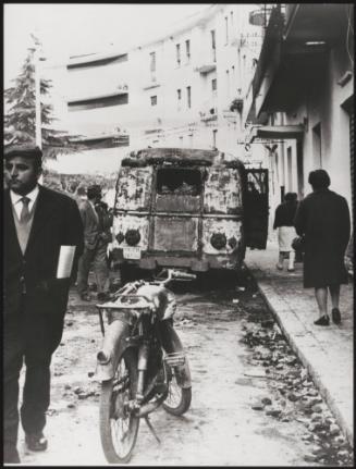 Pedestrians on a street pass a burnt out car, Battipaglia, 1969