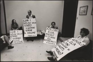 Gianfranco Spadaccia, the politician, stands with a protest placard against the newspaper 'Il Giorno' around his neck, with three other protesters from the Radical Party sitting with him