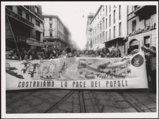 Milan 1982. A crowd of protesters march behind a large banner that reads 'costruiamo la pace dei popoli' down a street at a communist  peace march 