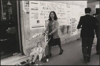 A woman walks down the street pushing a child in a pram; the wall behind her is plastered with protest posters attacking the police for its provocation and violence that led to the death of one of their activists