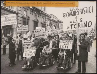 At a demonstration for the disabled in Rome, a group of people in the street, some in wheelchairs, are all holding protest placards