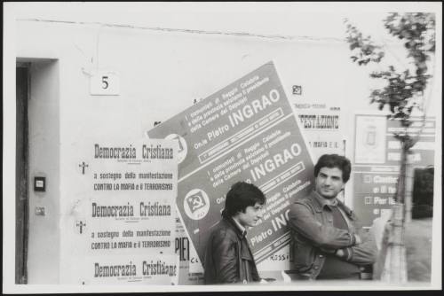 Two men lean against a wall bearing posters for the Communist and Democratic Parties during an anti Mafia demonstration in Calabria