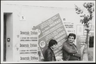 Two men lean against a wall bearing posters for the Communist and Democratic Parties during an anti Mafia demonstration in Calabria