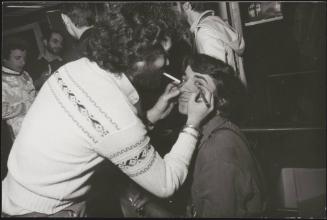 A man draws eyeliner on his friend 