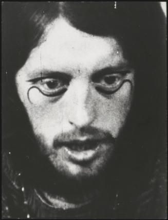 A close-up portrait of a man with long hair and black lines of make-up around his eyes