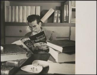 A man at a desk surrounded by lots of thick books, reading a Tempo annual magazine from 1951