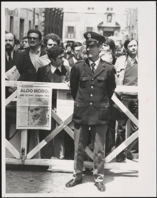 A policeman guards barriers with crowds behind him and the front page of the Christian Democracy newspaper, Il Popolo, bearing a large image of Aldo Moro pinned to it