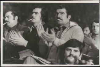 Photograph of crowd members at a political rally, focussed in on three men with moustaches, all smoking cigarettes