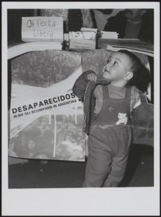 A toddler stands in front of a missing persons of Argentina sign 