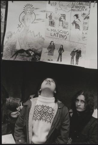 Two women at a feminist protest sit under a hand drawn poster criticising the 'Latin Male' 