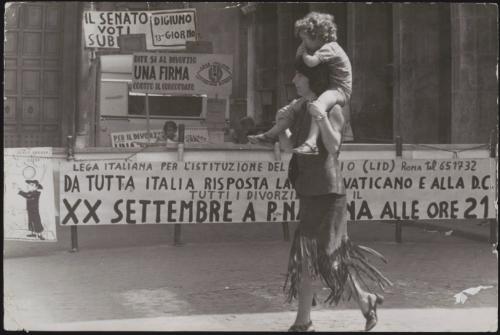 A mother carrying a child on her shoulders  walking past pro-divorce protest banners