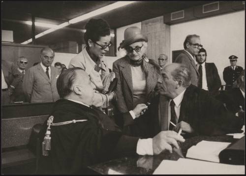 At the court case of Marino Vulcano, his mother, and the woman he fell for, Giuliana Meogrossi - former vice-director of the prison in which Vulcano had been serving a sentence for murder of his previous wife - listen intently to a discussion between the two seated men