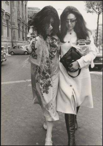 Two women with long hair, wearing long gowns and coats, walking arm in arm across an Italian car park