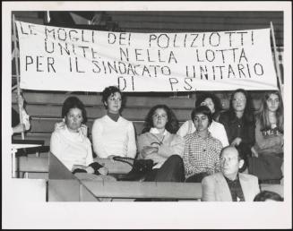 Policemen's wives taking part in a rally at the Palasport stadium, sitting under a banner reading "Le mogli dei poliziotti unite nella lotta per il sindacato unitario di P.S" [The wives of policemen joined in the fight for trade union unity of PS]
