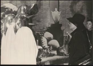 A woman in a hat and coat looking at a modern art sculpture made of white drapery and gleaming metal shields with protruding spikes
