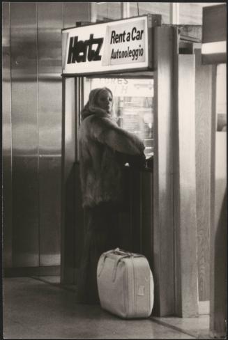 A woman in fur stands at the Hertz car rental kiosk in an airport 
