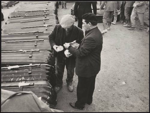 Two men sharing a sandwich beside a line of stacked coffins, for victims of the Belice earthquake