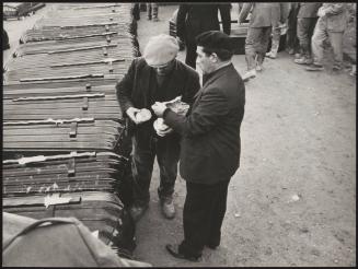 Two men sharing a sandwich beside a line of stacked coffins, for victims of the Belice earthquake