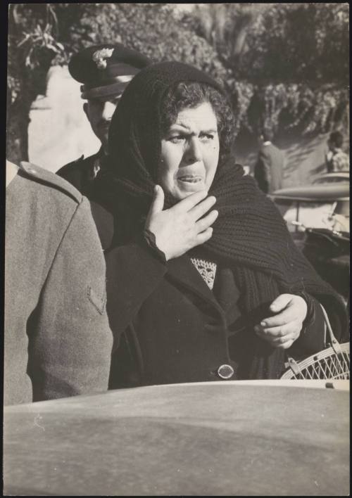 A survivor of the Belice earthquake wears a black shawl and carries a basket on a street between to policemen, Sicily, 1968