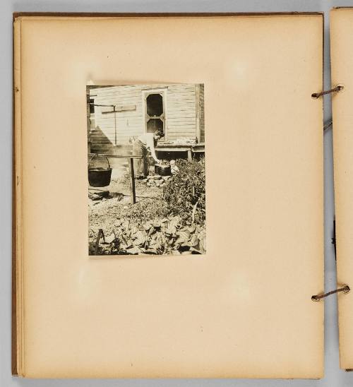 [A woman cooks in the yard with pots and a cauldron, a baby looks on] [Page 30 of Wells and Pots album]