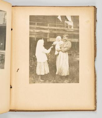 [Doukhobor woman holding a baby in her arms, as baby reaches out for a flower offered by a girl] [Page 75 of The Doukhobors II album]