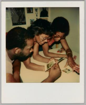 [Three people leaning over table and looking at polaroids]