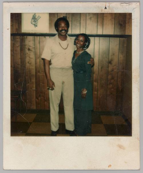R.Lee, 10-24-77 [Man with arm around woman standing in front of wood panelled wall]