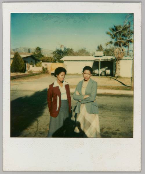 [Two women standing on sidewalk wearing long skirts, woman on right with arms crossed]
