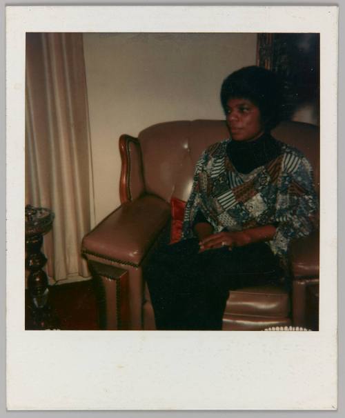 [Woman with afro sitting on brown loveseat wearing patterned patchwork top]
