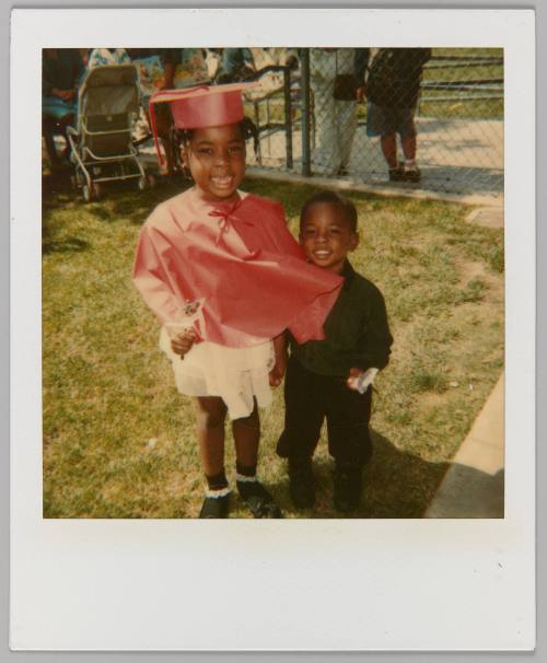 [Little girl wearing red graduation gown posing with little boy]
