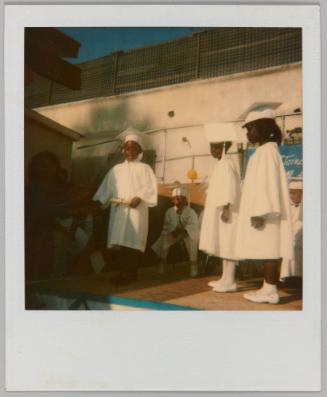 [Three children in white graduation gowns on stage]
