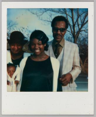 [Teenage girl in white cardigan posing with man in plaid suit and a woman holding up a baby, they are standing outside]