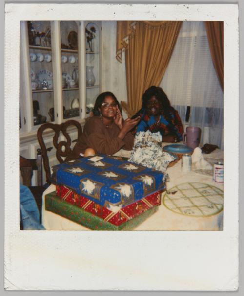 [Two women sitting at dining table, stack of Christmas presents in foreground]

