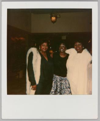 [Three women posing together, woman on left wearing white fur]
