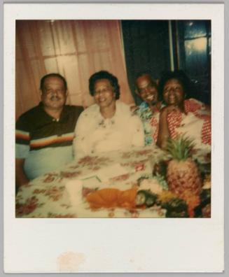 [Two men and two women sitting at table with floral tablecloth and fruit]
