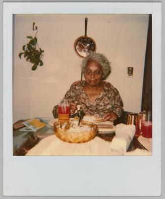 [Elderly woman eating a meal at the table, frying pan clock in background]
