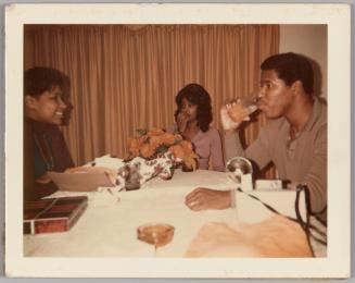 [Two women and a man sitting around dining table, man is drinking an orange beverage]
