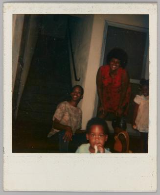 [Two women with little boy and girl smiling beside staircase]
