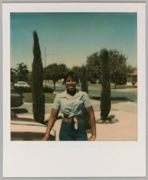 [Young woman in blue crop top standing in driveway with trees]
