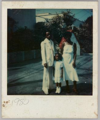1980 [Man, woman and child dressed in white outside with crucifix in background]
