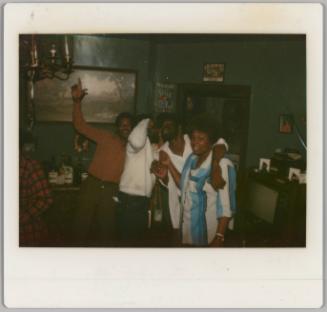 [Three men and a woman appear to be singing in home, man on left hold arm in the air]
