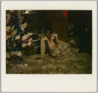 [Woman in plaid skirt laying on carpet next to Christmas tree]
