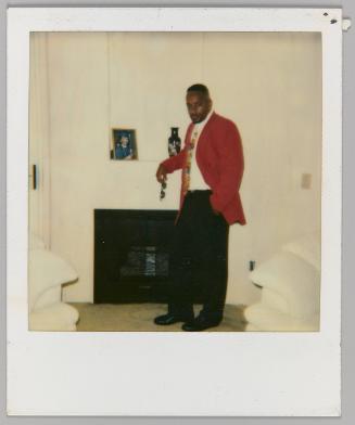 [Man in red blazer posing holding sunglasses in front of white fireplace]
