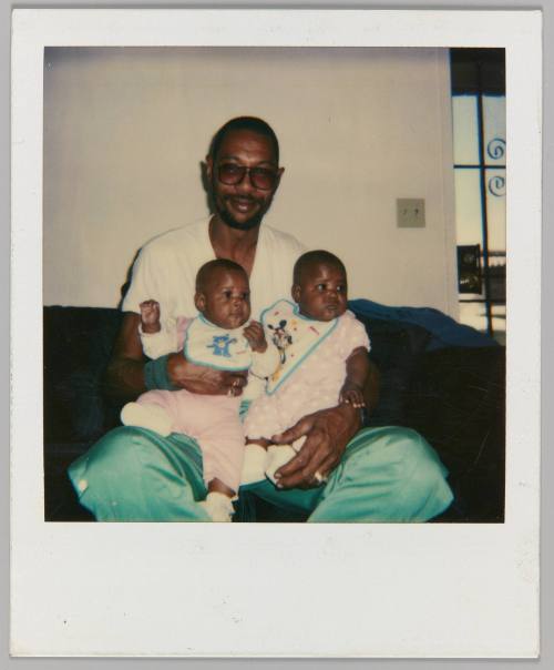 [Man in sunglasses seated on couch holding twin babies in lap]
