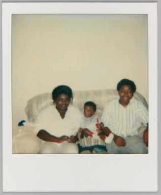 6/20/87 [Two women sitting on plastic wrapped couch smiling with baby between them]
